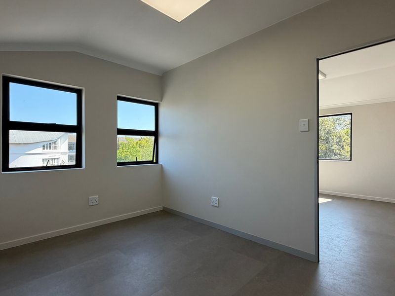 A bright, empty office space featuring neutral walls, tiled flooring, and large windows providing natural light, with an adjacent room visible through the doorway.