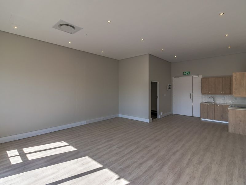 A bright, modern open-plan commercial space featuring light wood-look laminate flooring, recessed lighting, and a compact kitchenette with built-in cabinetry and sink.