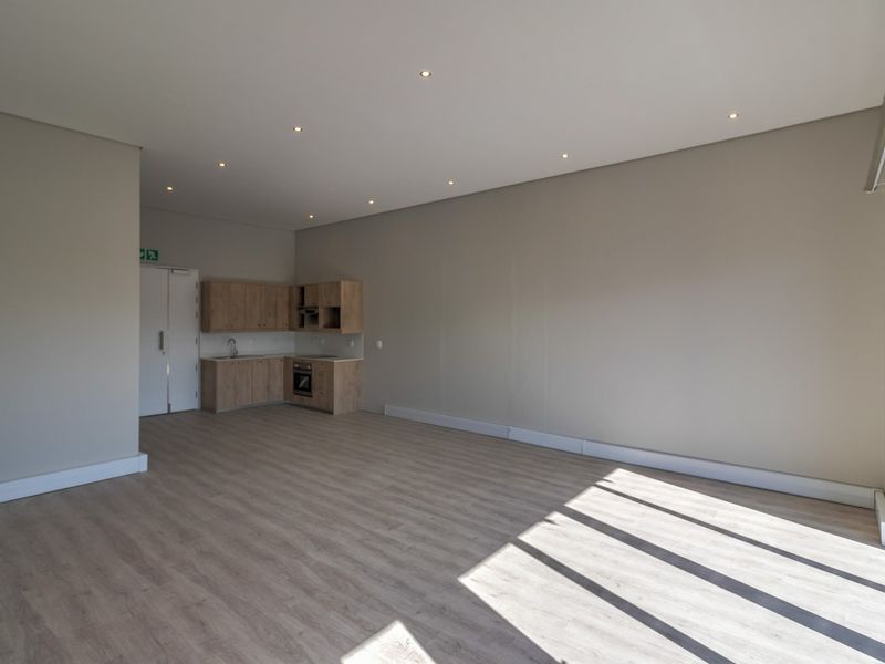 A bright, modern open-plan commercial space featuring light wood-look laminate flooring, recessed lighting, and a compact kitchenette with built-in cabinetry and appliances.