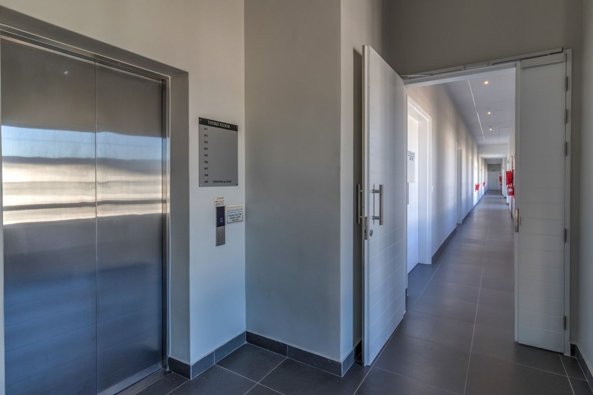 A clean, modern commercial hallway featuring tiled floors, a stainless steel elevator, and recessed lighting leading to multiple office units.