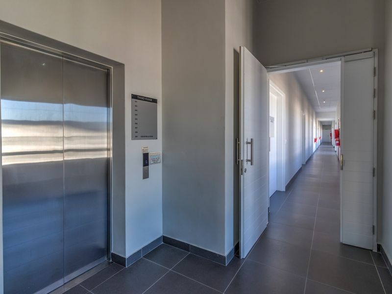 A clean, modern commercial hallway featuring tiled floors, a stainless steel elevator, and recessed lighting leading to multiple office units.