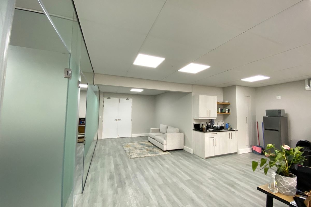 Modern open-plan office space featuring a glass partition, recessed lighting, laminate flooring, and a compact kitchenette area with built-in cabinetry.