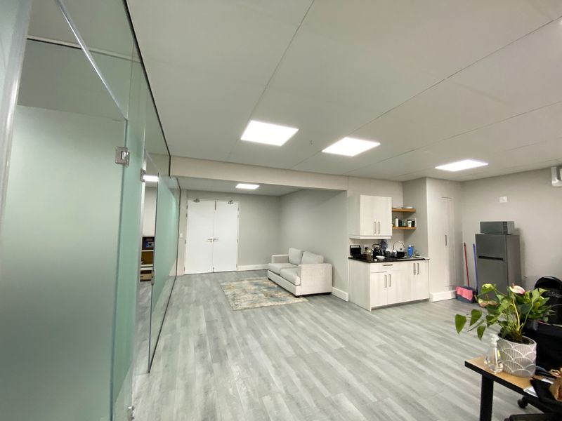 Modern open-plan office space featuring a glass partition, recessed lighting, laminate flooring, and a compact kitchenette area with built-in cabinetry.