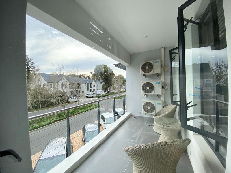 A modern covered balcony featuring a glass balustrade, tiled flooring, and multiple wall-mounted air conditioning units, overlooking the street and surrounding office complex.