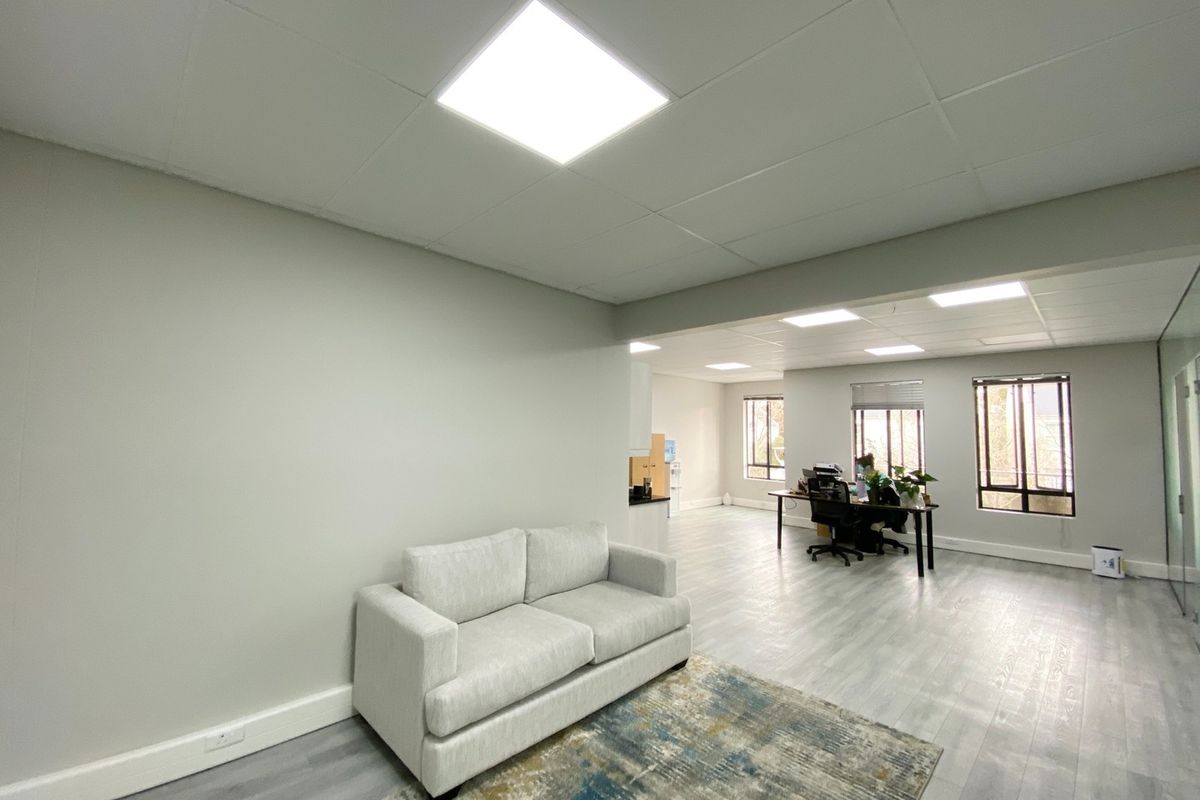 A bright, open-plan office space featuring modern laminate flooring, suspended ceilings with recessed LED lighting, and large windows providing ample natural light.
