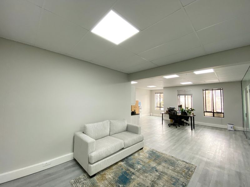 A bright, open-plan office space featuring modern laminate flooring, suspended ceilings with recessed LED lighting, and large windows providing ample natural light.