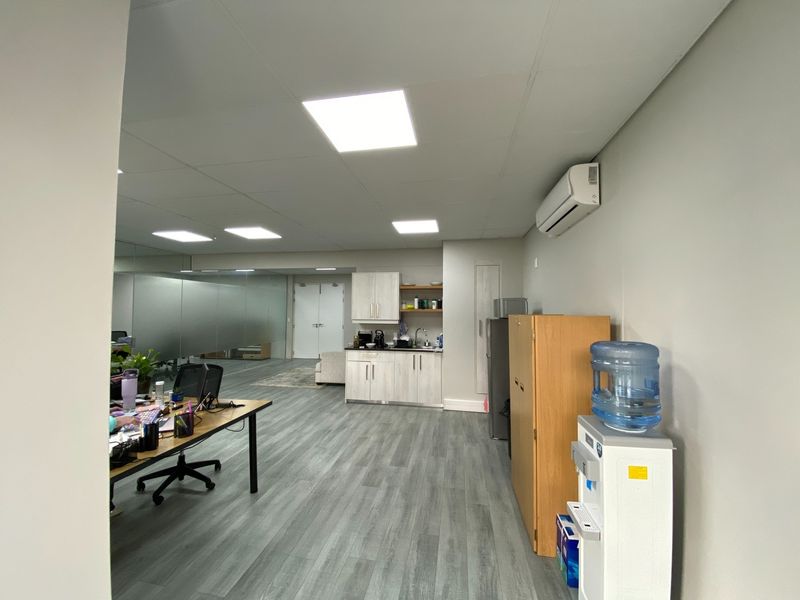 A modern office space featuring a functional kitchenette, laminate flooring, recessed lighting, and a mirrored wall, providing a clean and professional work environment.
