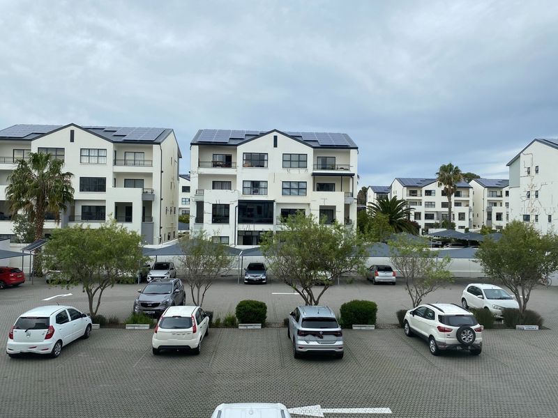 Elevated view overlooking a paved parking lot with shade cloth carports and multi-story office or residential buildings with rooftop solar panels.