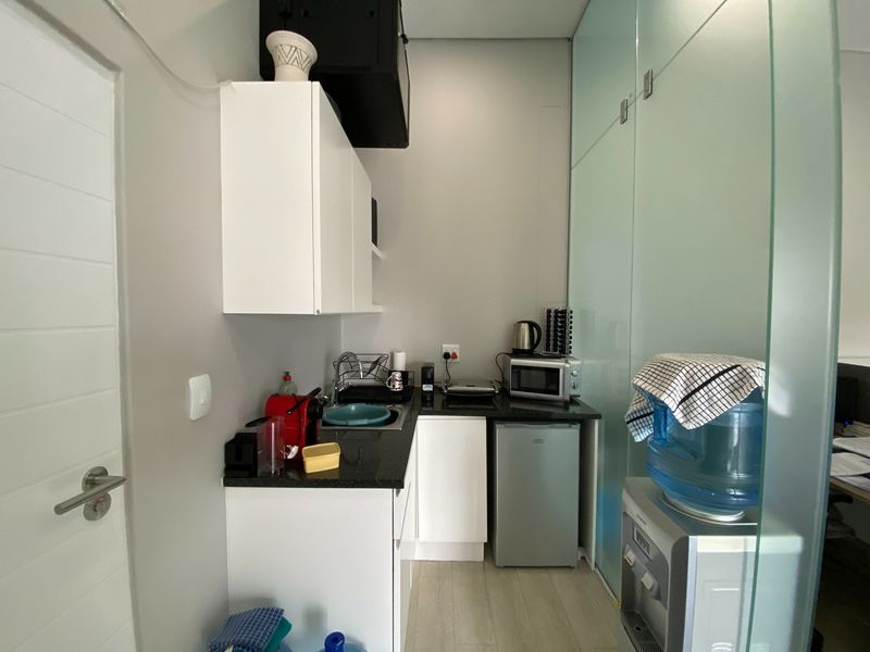 A compact office kitchenette featuring white cabinetry, granite countertops, a stainless steel sink, and a modern glass partition wall.
