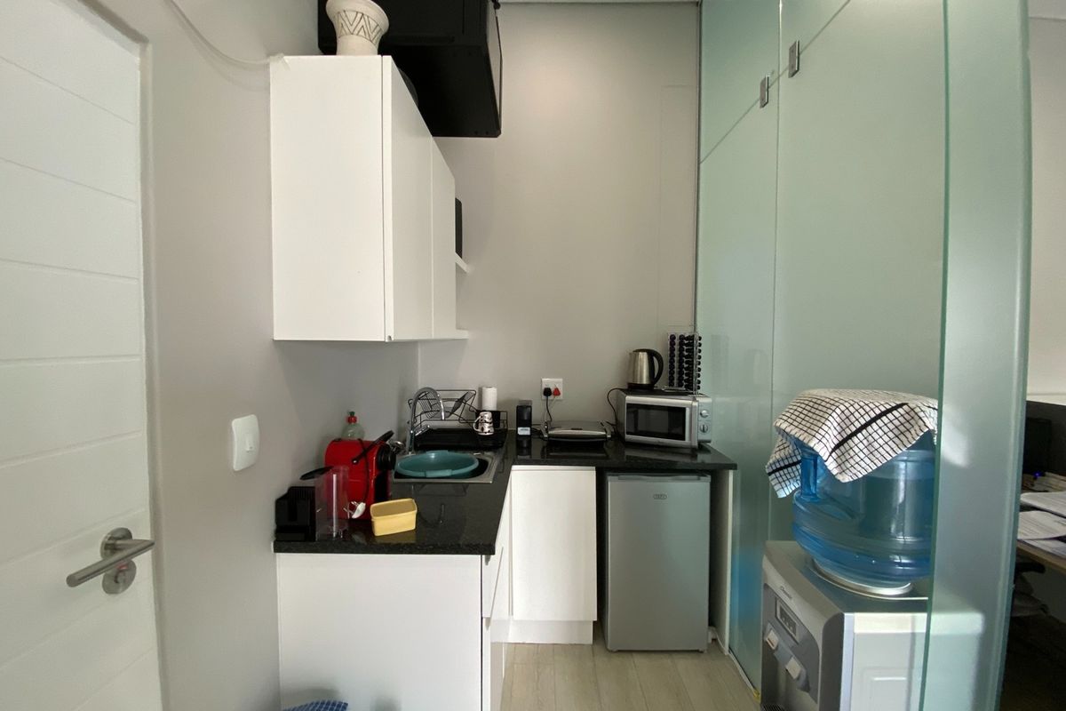 A compact office kitchenette featuring white cabinetry, granite countertops, a stainless steel sink, and a modern glass partition wall.