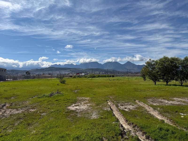 A wide-angle view of a vacant commercial land plot featuring natural grassland, a perimeter boundary wall, and scenic mountain views in the background.
