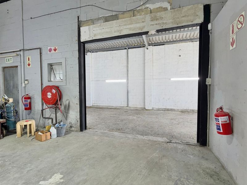 Industrial utility space featuring concrete flooring, exposed brick walls, fire safety equipment, and a large access opening to an adjacent warehouse area.