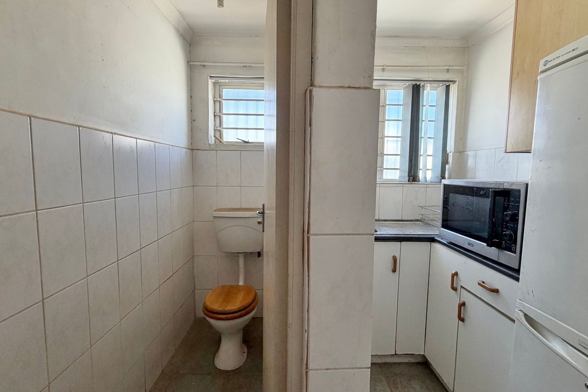 A compact industrial kitchen area featuring built-in cabinetry and an adjacent private toilet facility with tiled walls and security window bars.