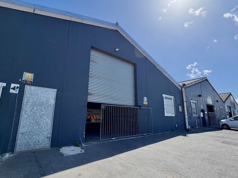 Front elevation of an industrial warehouse unit featuring a large roller shutter door, security gate, and paved driveway access.