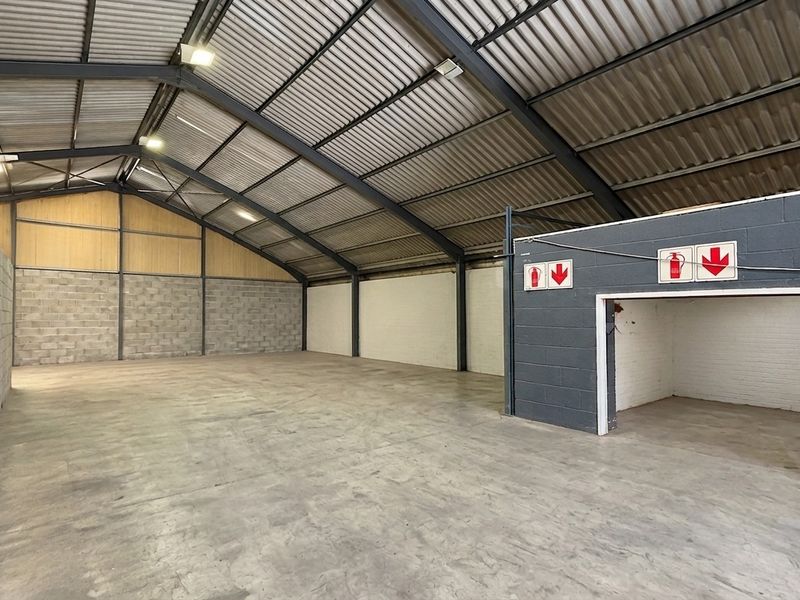 Spacious industrial warehouse unit featuring exposed steel portal framing, concrete flooring, and high-clearance corrugated metal roofing with integrated fire safety signage.
