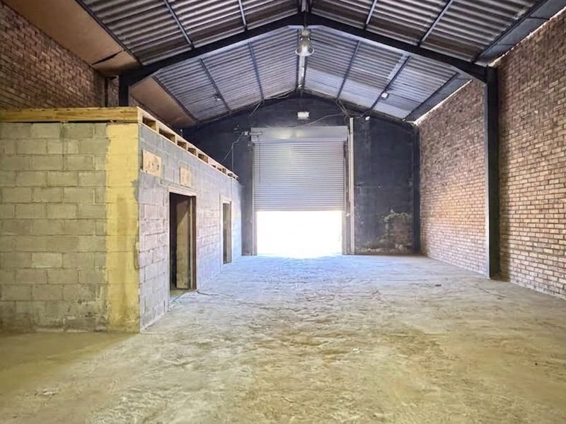 Spacious industrial warehouse unit featuring exposed brick walls, high-clearance steel portal frame, concrete flooring, and a large roller shutter door for vehicle access.