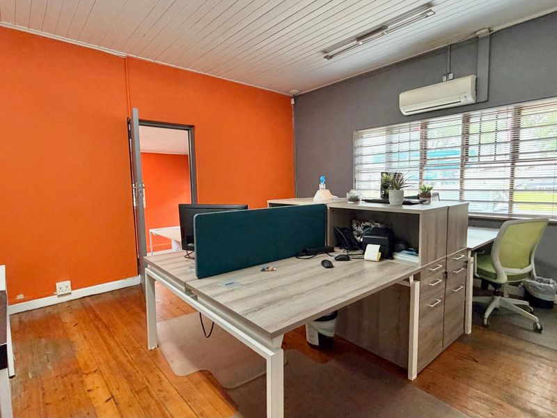 A functional office space featuring timber flooring, an air conditioning unit, and large windows with venetian blinds, offering a practical workspace environment.