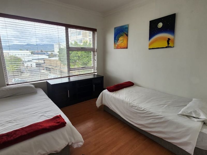 A bedroom featuring laminate flooring and large windows with venetian blinds, offering natural light and an urban view.