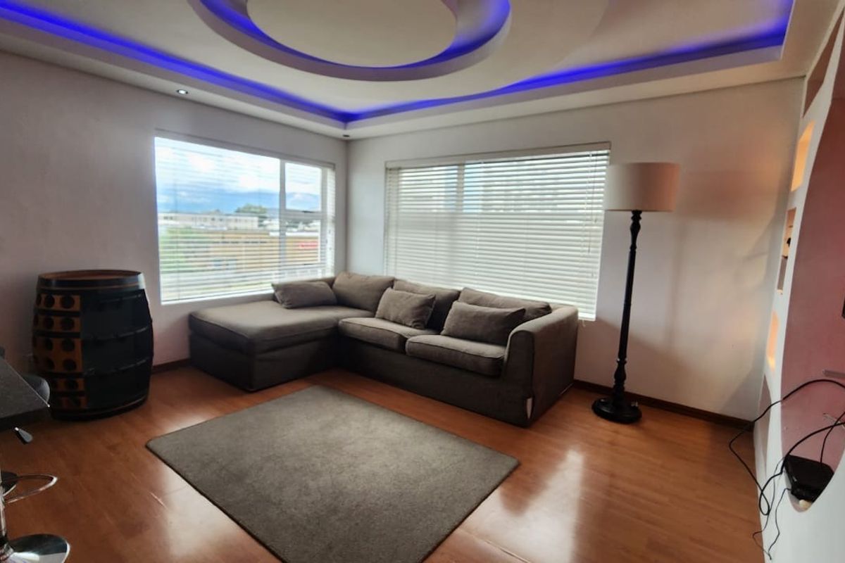 A modern living room featuring laminate flooring, large windows with venetian blinds, and a distinctive recessed ceiling with integrated blue LED accent lighting.