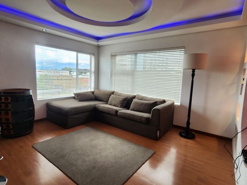 A modern living room featuring laminate flooring, large windows with venetian blinds, and a distinctive recessed ceiling with integrated blue LED accent lighting.