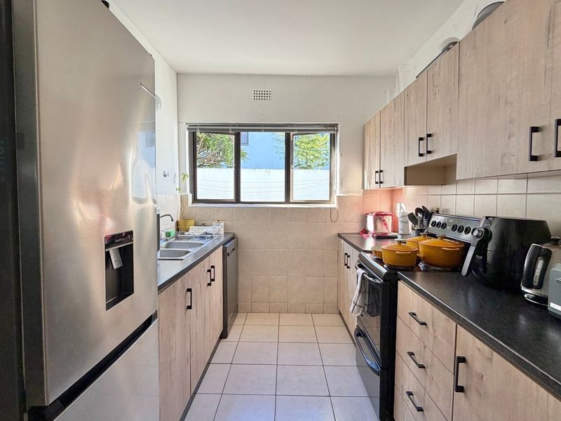 A functional galley-style kitchen featuring light wood-look cabinetry, dark countertops, tiled backsplash, and tiled flooring with a window providing natural light.