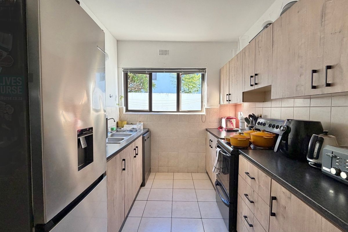 A functional galley-style kitchen featuring light wood-look cabinetry, dark countertops, tiled backsplash, and tiled flooring with a window providing natural light.