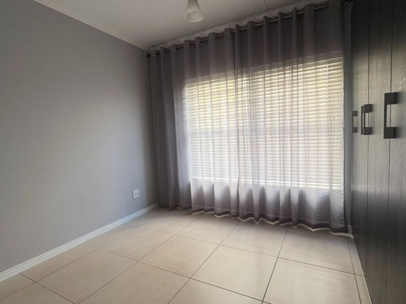 A clean, empty bedroom featuring neutral tiled flooring, built-in cupboards, and window blinds, offering a versatile space ready for personalization.