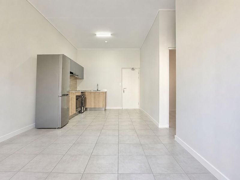 A bright, open-plan living and kitchen area featuring neutral tiled flooring, modern cabinetry, and integrated kitchen appliances in a clean, minimalist space.
