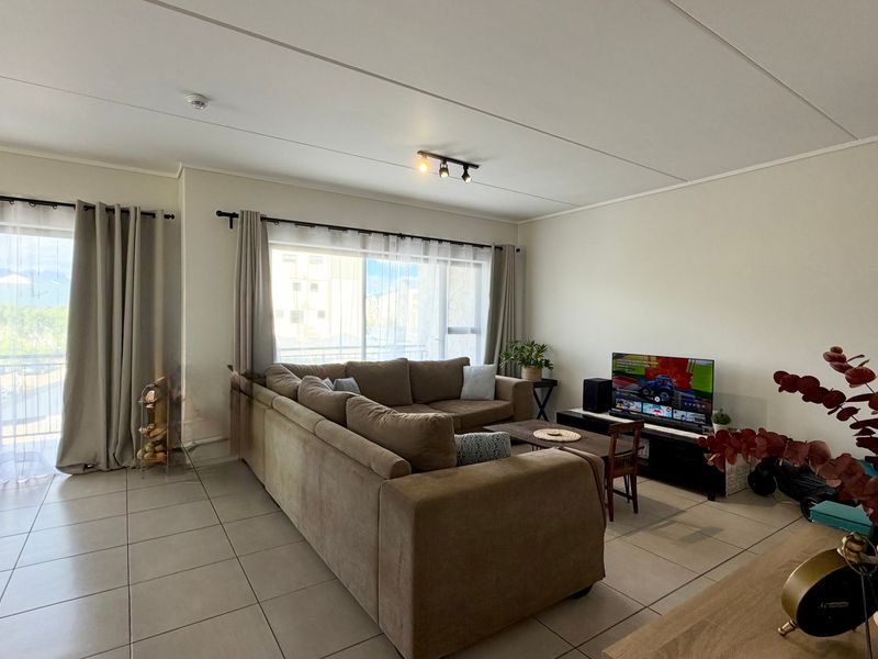 A bright and spacious living room featuring neutral tiled flooring, modern track lighting, and a large sliding glass door providing access to the balcony.