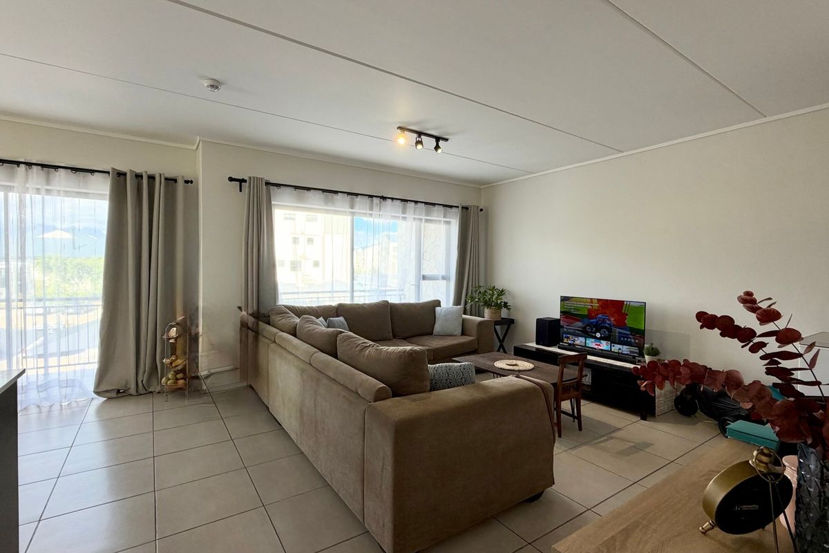 A bright and spacious living room featuring neutral tiled flooring, modern track lighting, and a large sliding glass door providing access to the balcony.