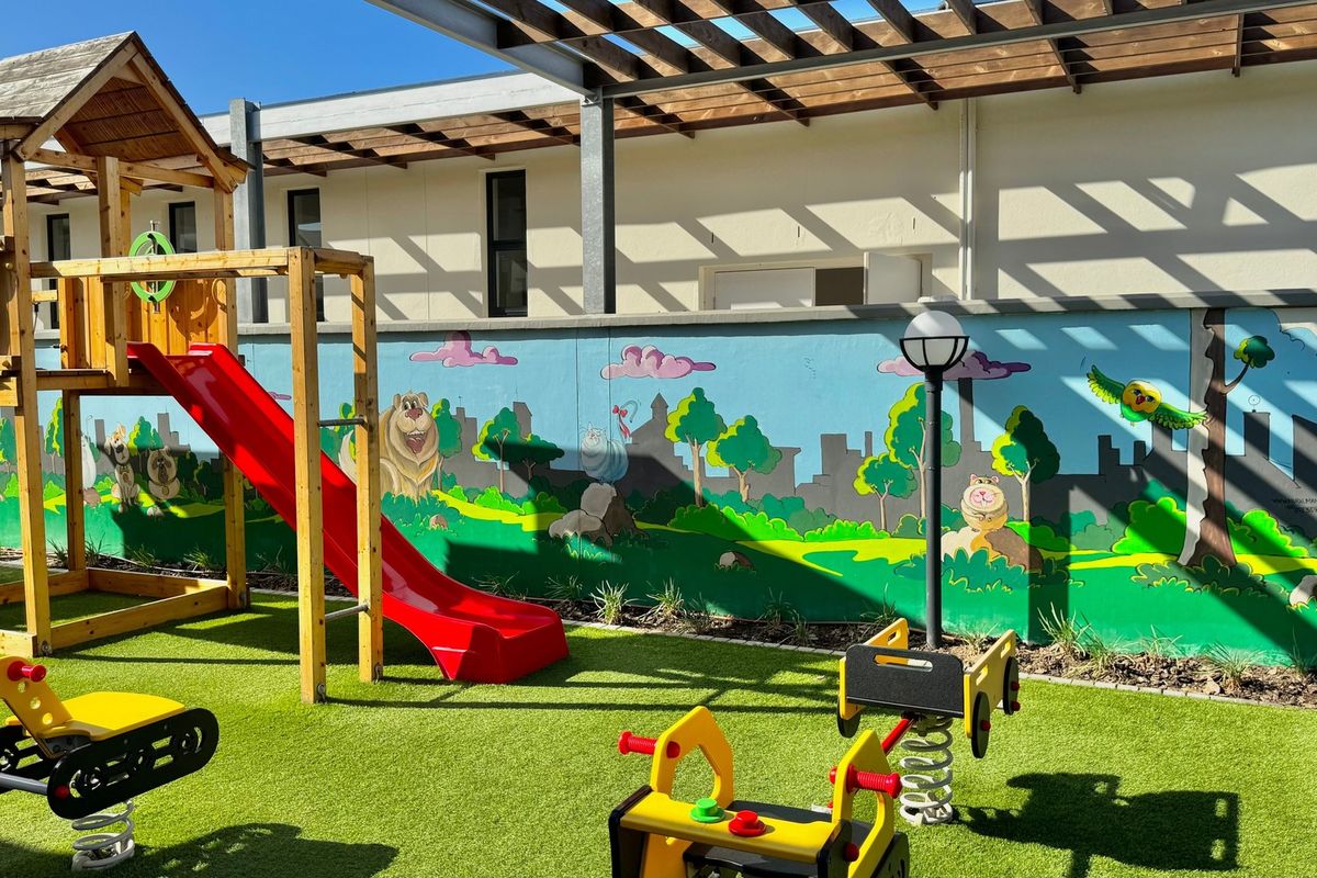 A well-maintained outdoor playground area featuring artificial grass, a wooden pergola, and a decorative mural on the boundary wall.