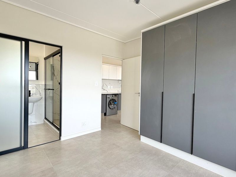 A modern, empty bedroom featuring sleek grey built-in cupboards, neutral tiled flooring, and direct access to an en-suite bathroom.