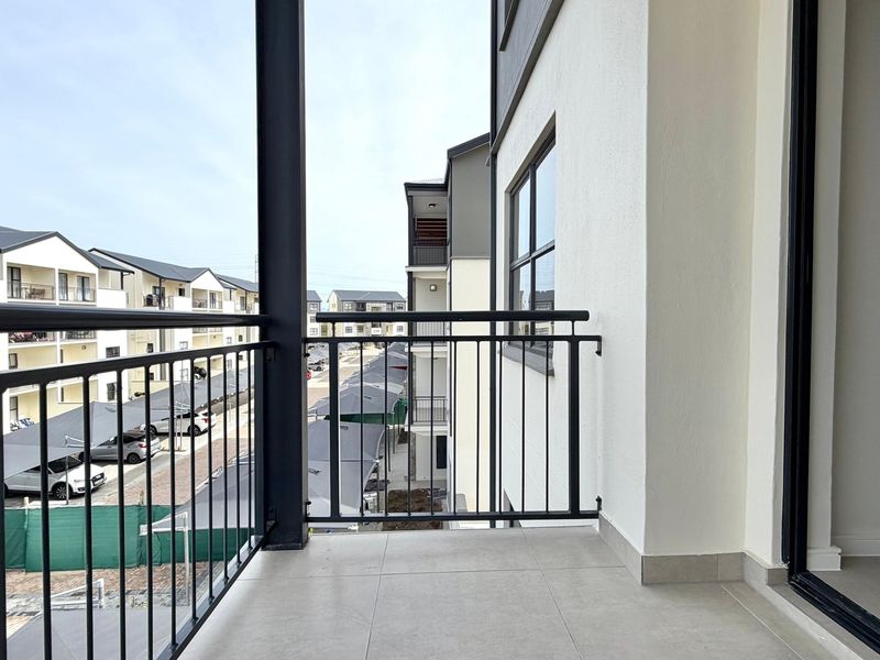 A clean, modern balcony featuring tiled flooring and a black metal balustrade, overlooking the apartment complex parking area.