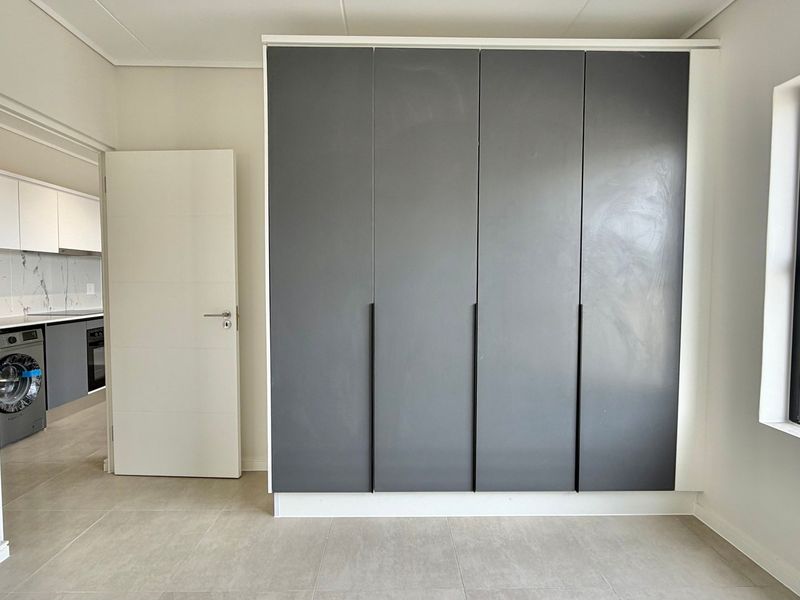 A bright, modern bedroom featuring neutral tiled flooring, a large built-in wardrobe with dark grey doors, and a window with natural light.