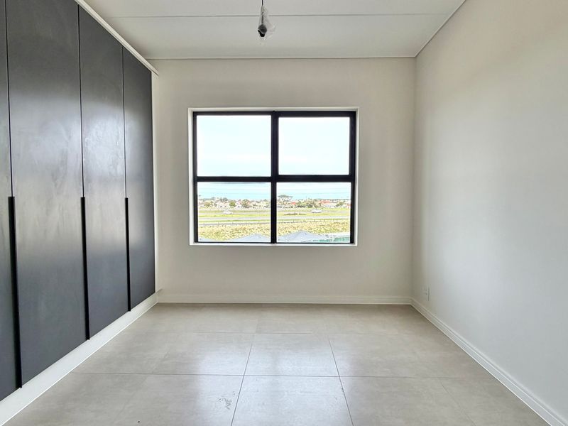 A bright, empty bedroom featuring modern dark built-in cupboards, neutral tiled flooring, and a large window with scenic exterior views.