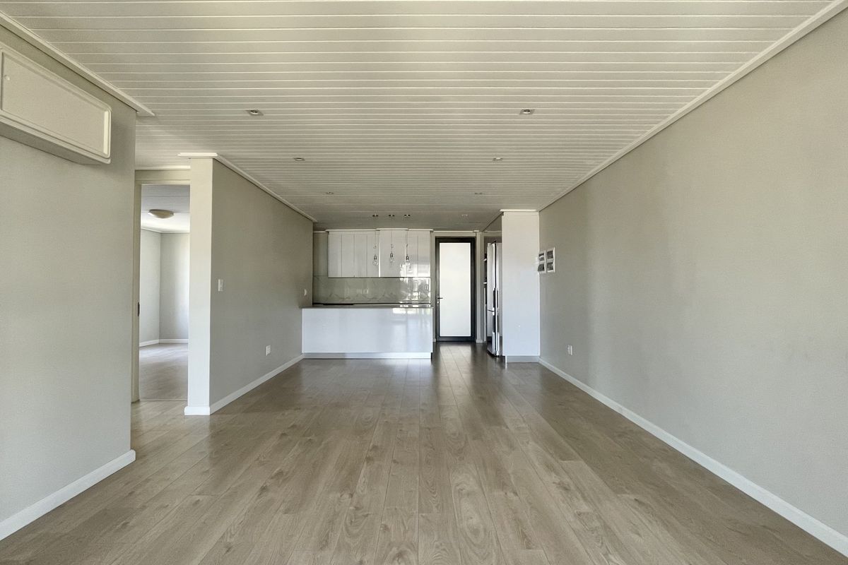 Spacious open-plan living area featuring light laminate flooring, recessed lighting, a wall-mounted air conditioning unit, and a modern kitchen with white cabinetry.