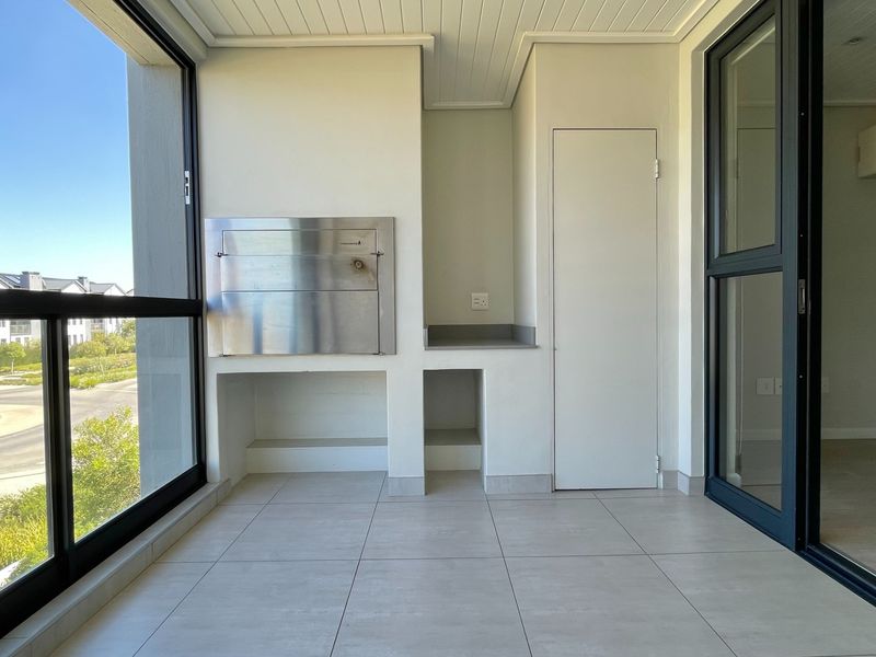 A modern covered balcony featuring a built-in stainless steel braai, tiled flooring, and large sliding glass doors providing ample natural light.