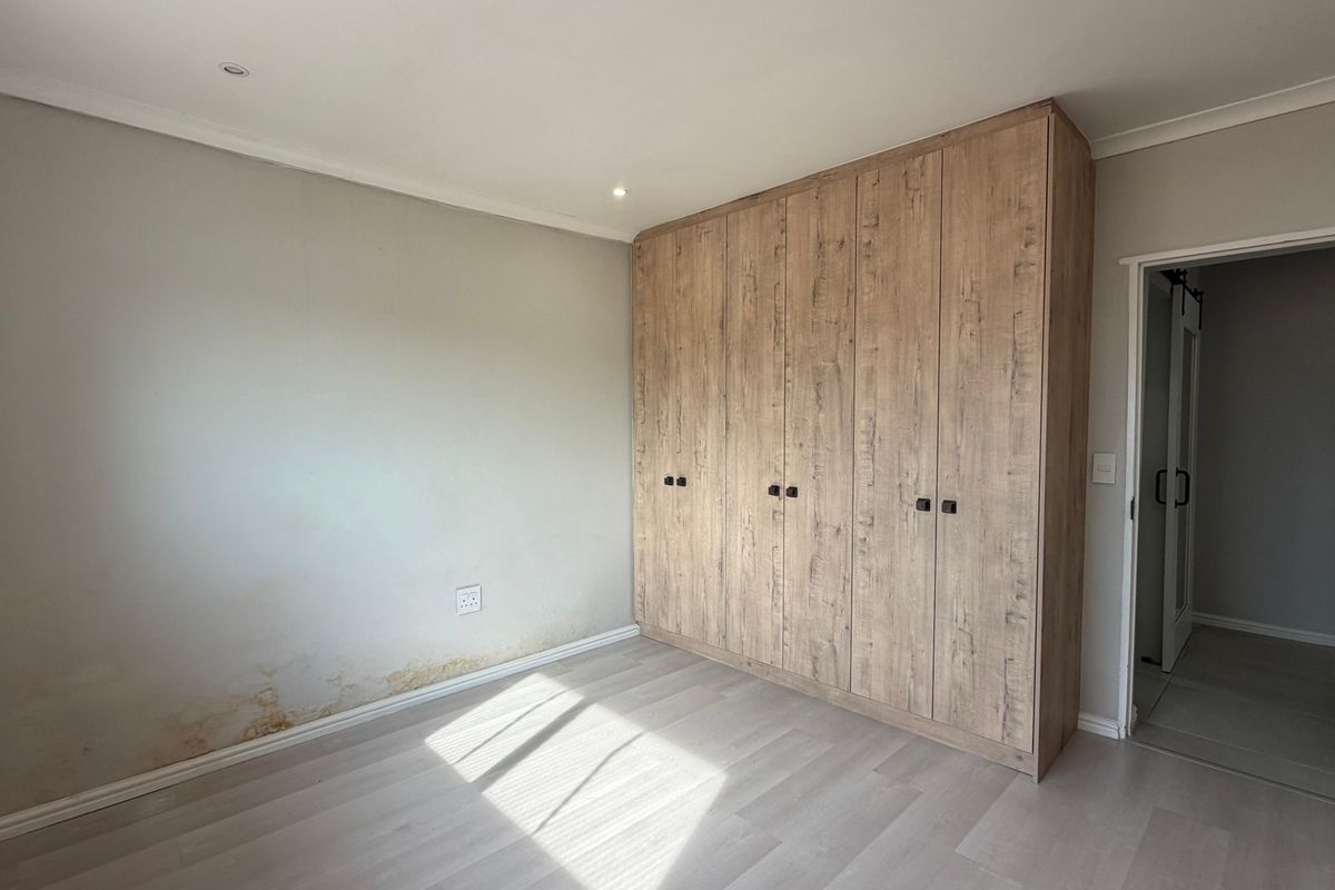 A bedroom featuring light laminate flooring and a large built-in wooden wardrobe, showing signs of moisture damage on the lower wall section.