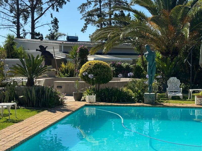 A well-maintained outdoor pool area featuring a clear swimming pool with brick coping, surrounded by established garden landscaping and mature trees.