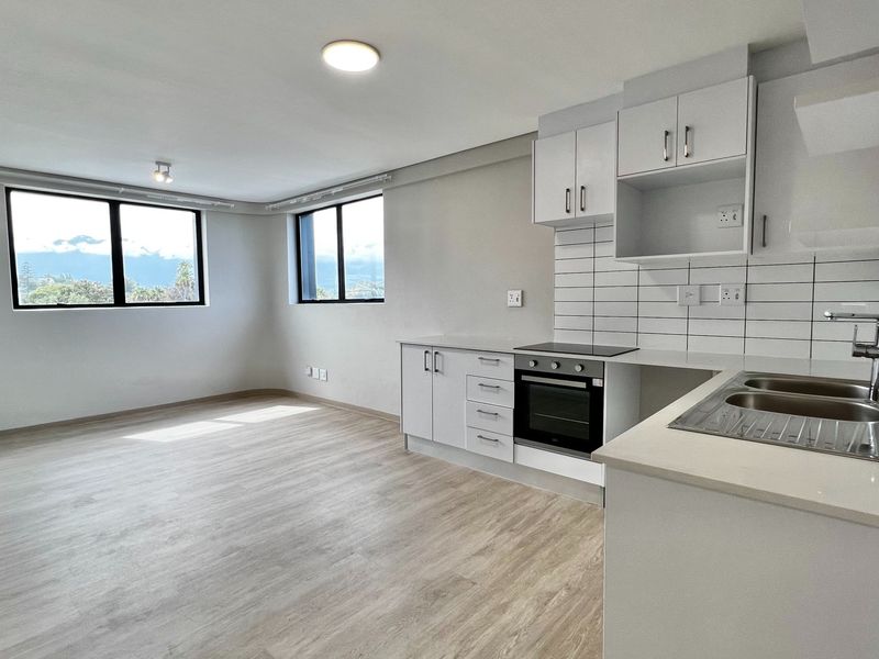Bright open-plan living area featuring a modern kitchen with built-in oven, hob, and sleek cabinetry, complemented by light laminate flooring and large windows.