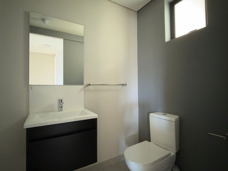 A clean and modern bathroom featuring a vanity cabinet with basin, wall-mounted mirror, toilet, and a frosted window for natural light.