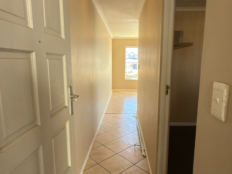 A narrow hallway with tiled flooring and neutral walls, leading towards a windowed area. The space is vacant and requires minor cosmetic updates.