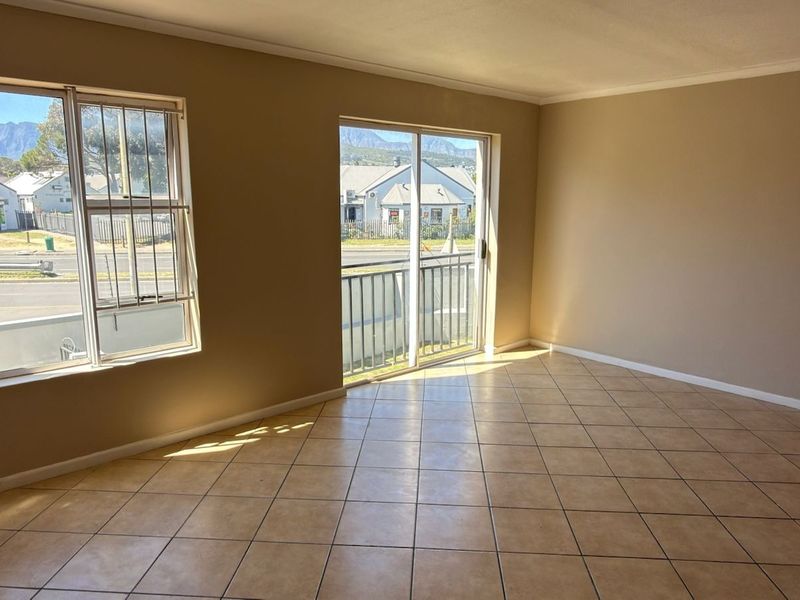 A bright, empty living room featuring neutral tiled flooring, large windows with security bars, and a sliding glass door leading to a balcony.