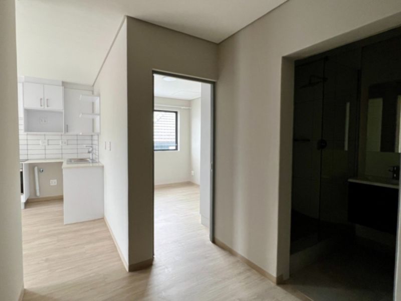 An open-plan apartment interior featuring light laminate flooring, a modern kitchen with white cabinetry, and access to a bedroom and bathroom.