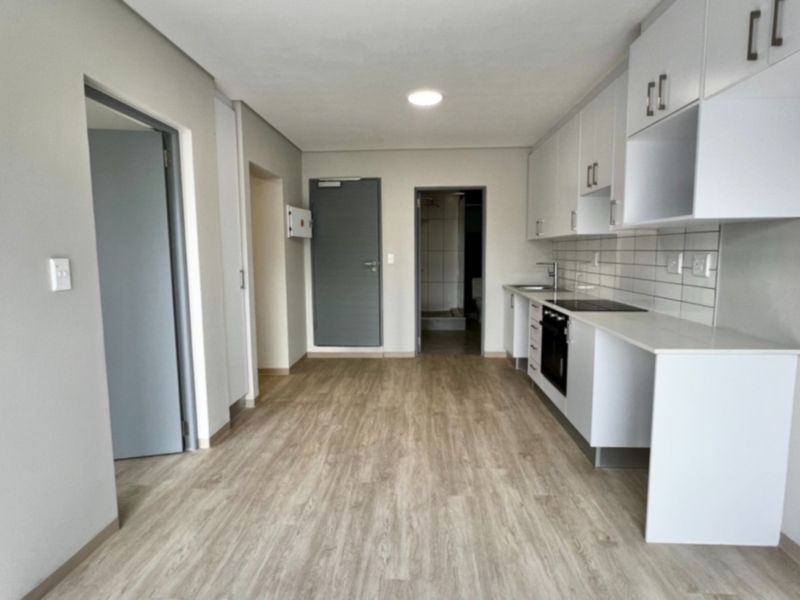 Modern open-plan kitchen featuring sleek white cabinetry, subway tile backsplash, built-in oven and hob, and light wood-look laminate flooring in a clean, vacant space.