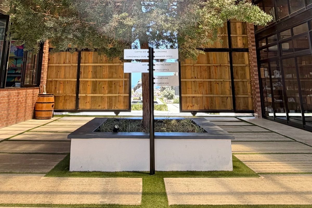 A modern, well-maintained courtyard area featuring a central planter with a tree, paved walkways, and directional signage for building amenities.