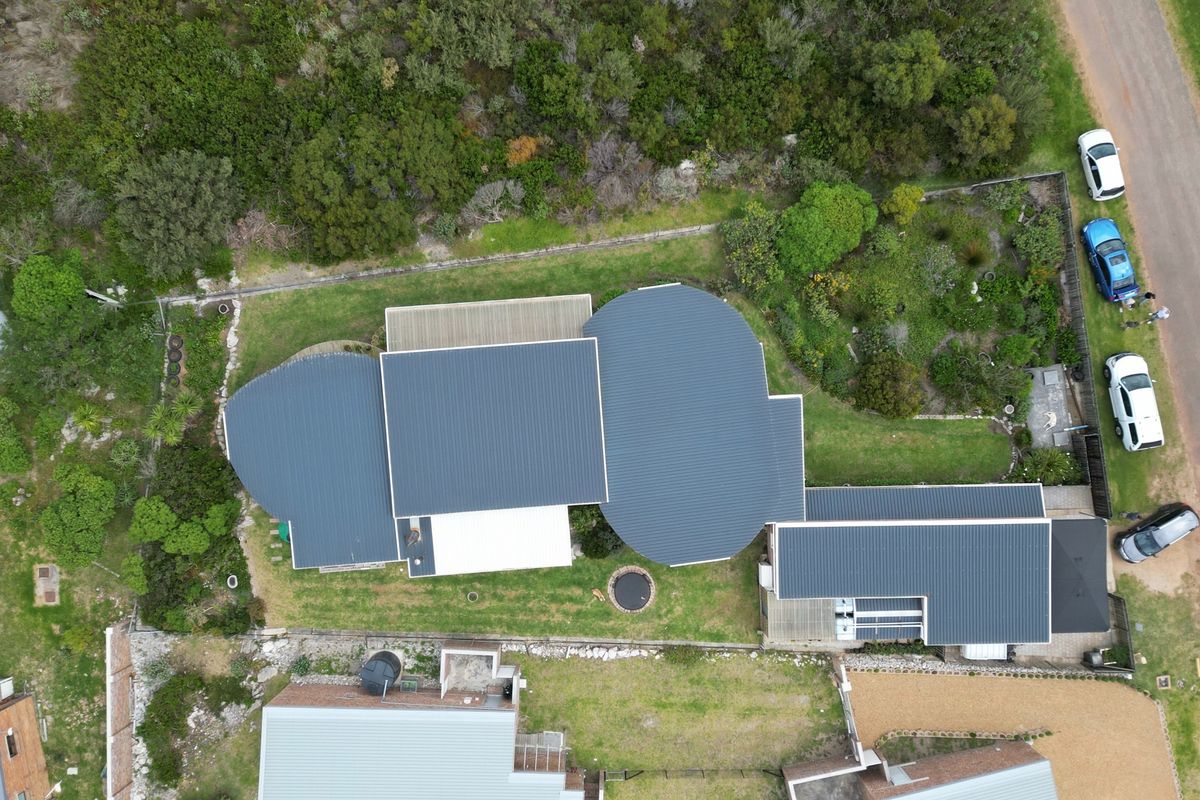 Aerial view of the property showcasing the unique curved metal roof design, integrated solar panels, timber decking, and surrounding landscaped garden areas.