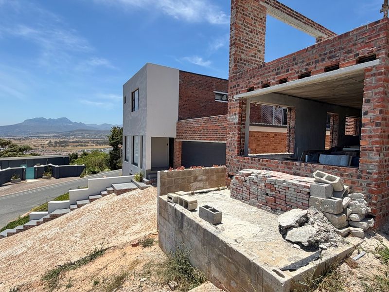 A property under construction featuring exposed brickwork and concrete structural elements, situated on a sloped lot with views of the surrounding area.