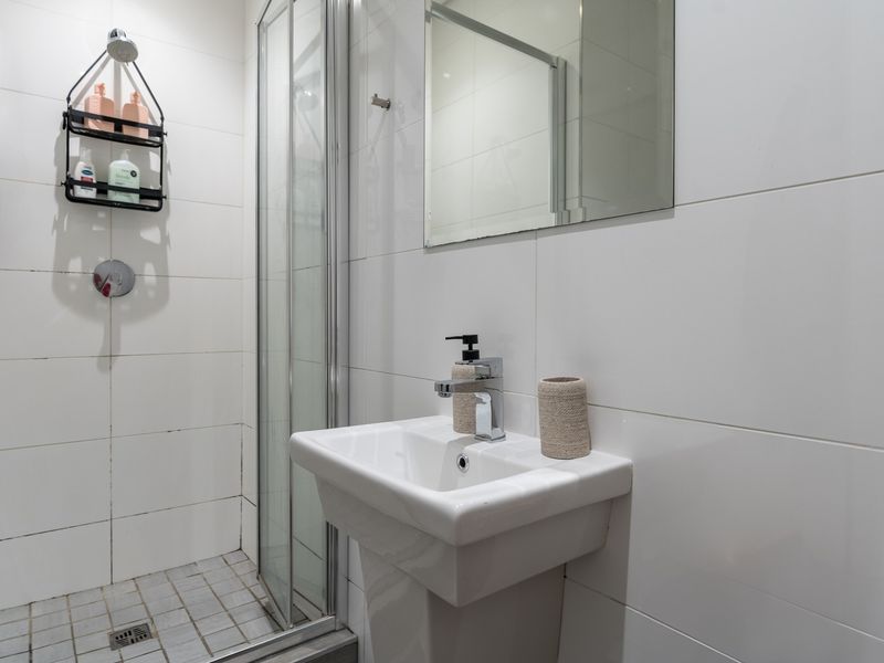 A clean, modern bathroom featuring a pedestal washbasin, chrome fixtures, a glass-enclosed shower, and neutral wall tiling.