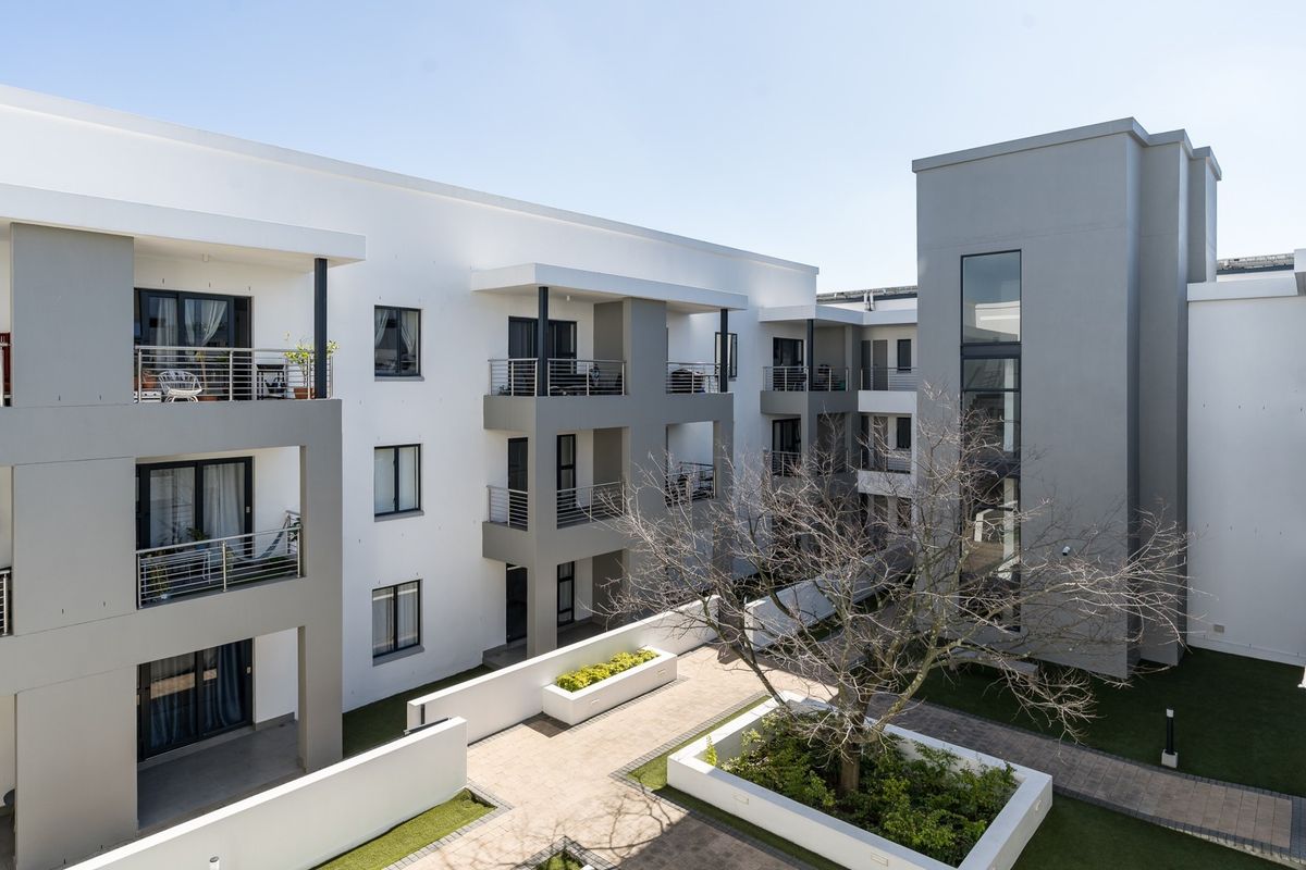 A modern apartment complex featuring a central paved courtyard, raised garden beds, and private balconies with stainless steel railings.